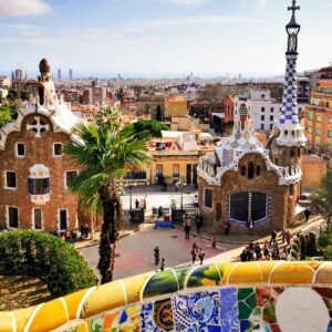 A colorful, panoramic view of Park Güell's iconic entrance with Barcelona cityscape in the background.